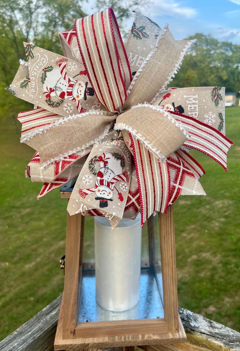 May include: A wooden lantern with a white candle inside. The lantern is decorated with a large burlap and red and white striped bow with snowman and Christmas themed fabric accents. The bow has the words "Merry Christmas" printed on it.