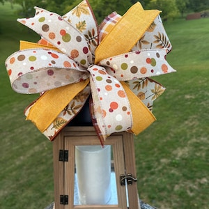 May include: A decorative lantern with a large bow on top. The bow features ribbons with fall-themed patterns, including polka dots, leaves, and plaid. The lantern is made of wood and has a glass panel, with a white candle inside.