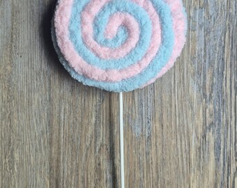 May include: A large, plush lollipop with a spiral design in alternating light blue and pink. The candy is mounted on a white stick, set against a wooden background. The lollipop is a whimsical decorative item.