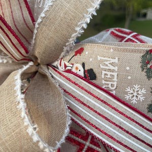 May include: A close-up of a Christmas bow made with burlap ribbon, red and white striped ribbon, and plaid ribbon. The bow has a frayed edge and features the word "Merry" printed on the ribbon.