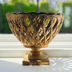 May include: A gold-coloured pedestal bowl featuring a textured, lattice-style openwork design. The bowl has a scalloped rim and sits on a square base, with a rough, metallic finish that creates a distinct pattern of light and shadow.