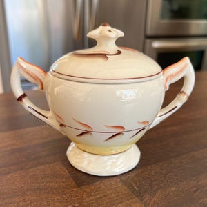 May include: Cream-colored ceramic sugar bowl with a lid and two handles. The bowl has brown and orange accents, with a yellow base. The lid is topped with a decorative finial. A vintage kitchenware item.