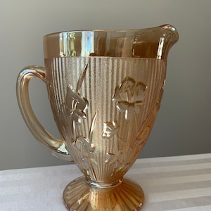 May include: A vintage amber glass pitcher with a floral design and a ribbed pattern. The pitcher has a wide base and a handle.