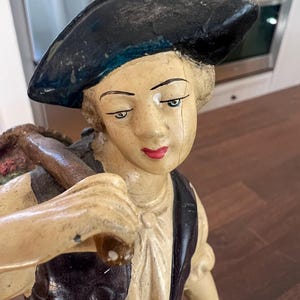 May include: A vintage figurine of a person wearing a black beret and vest. The figurine has a cream-colored face with red lips and green eyes. The figure is carrying a bundle over their shoulder.