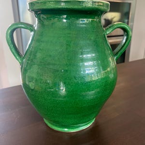 May include: A vibrant green ceramic vase with two curved handles. The vase has a bulbous body and a wide, slightly flared rim. The glossy finish reflects light, highlighting the texture of the ceramic. A decorative piece for home decor.