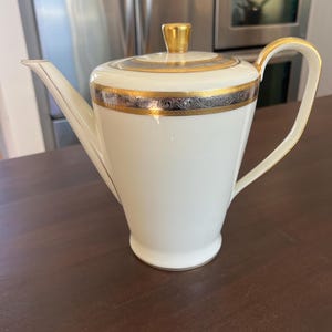 May include: A white porcelain teapot with gold trim and a gold handle. The lid has a gold knob and is also trimmed with gold. The teapot has a decorative band of silver and black near the top. The teapot is on a dark brown wooden surface.