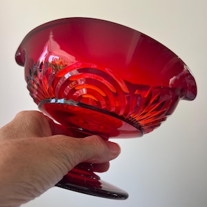 May include: A ruby red glass bowl with a pedestal base. The bowl has a wide, curved rim and is decorated with a repeating fan and swirl pattern. The bowl is held in a hand against a white background.
