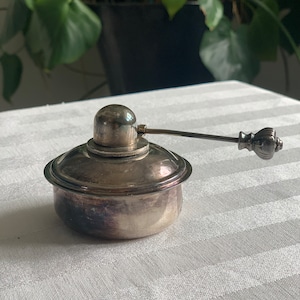 May include: A silver-toned metal inkwell featuring a rounded base, a domed lid with a central knob, and a long, slender horizontal arm extending to the right, ending in a decorative, fluted, bulbous handle. The metal surface shows signs of tarnish.