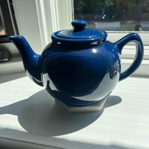 May include: A glossy, cobalt blue ceramic teapot with a matching lid and handle. The teapot sits on a white, hexagonal base, and is positioned on a white windowsill. The teapot is designed for brewing tea.