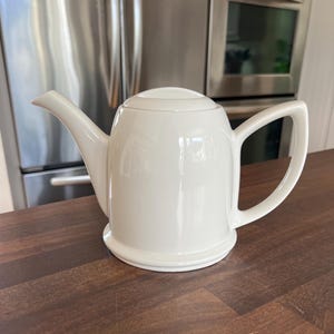 May include: A cream-coloured ceramic teapot with a lid, spout, and handle. The teapot is on a dark brown wooden surface. The background features a stainless steel appliance and a white wall.