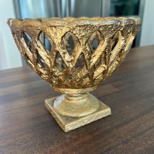 May include: A gold metal decorative bowl with a square base and an intricate openwork design. The bowl is made of cast iron and has a distressed finish.