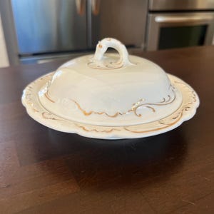 May include: A white ceramic butter dish with a domed lid and a matching base. The piece features a scalloped edge and delicate gold filigree scrollwork patterns along the rim of the base and the base of the lid, topped with a curved handle.