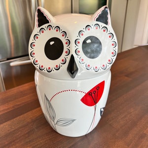 May include: White ceramic owl cookie jar with black and red details. The owl has large black eyes and a floral design on its body.