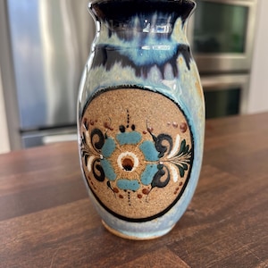 May include: A ceramic vase with a blue and tan glaze. The vase features a central circular design with a turquoise floral pattern and brown accents. The top rim is dark blue.