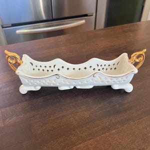 May include: A white porcelain serving dish with a scalloped edge and pierced design. The dish has gold-coloured handles and trim, and rests on four small feet. This decorative piece is ideal for serving snacks or displaying small items.