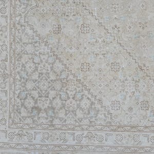 May include: A vintage rug with a faded, light beige background and a geometric pattern in shades of gray and blue. The rug has a border with a repeating design.