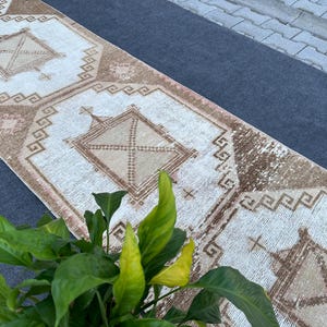 May include: A long, rectangular runner rug featuring a repeating pattern of large, light-colored hexagonal medallions outlined in brown. Each medallion contains a central geometric diamond shape with intricate cross-hatching and small cross motifs.