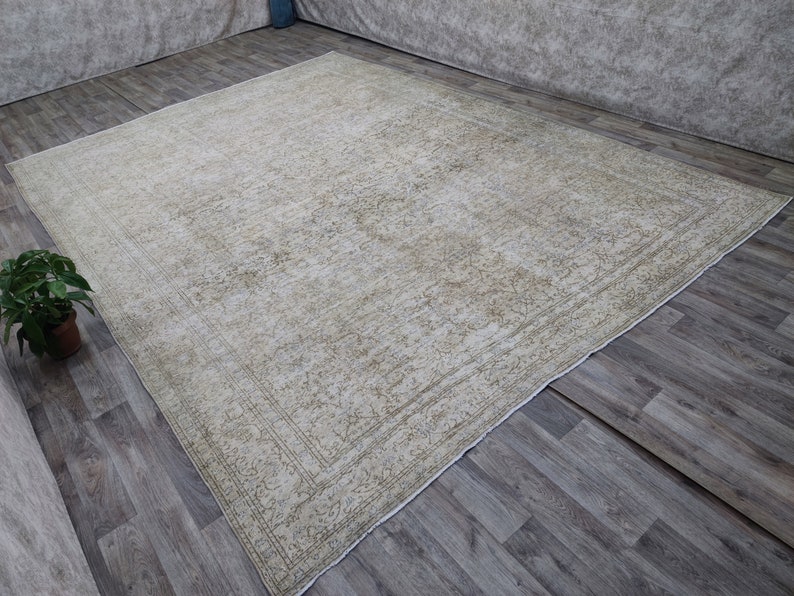 May include: A vintage cream-colored rug with a subtle floral pattern and a border. The rug is laid out on a wooden floor.
