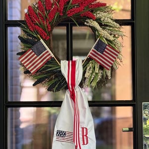 May include: A patriotic wreath with red, white, and blue elements. The wreath features red floral accents, greenery, and small American flags. A white fabric banner with red stripes and an embroidered American flag and the letter "B" hangs below.