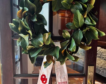 Op de afbeelding: Een grote, cirkelvormige krans van groene magnoliabladeren met bruine en gele accenten hangt aan een houten deur. Een wit lint met de tekst "BOOMER SOONER" en een rood logo is aan de krans bevestigd.