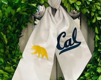 Op de afbeelding: Een witte stoffen krans met een gouden beer en het woord "Cal" in blauwe letters. De krans is versierd met groene buxus.