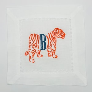Könnte beinhalten: Quadratische weiße Cocktail-Serviette aus Leinen mit einem gestickten orangefarbenen Tigerdesign und dem marineblauen Buchstaben "B". Die Serviette hat eine genähte Bordüre.