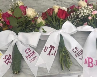 Puede incluir: Ramos de rosas de varios colores, incluyendo amarillo, rosa y rojo, atados con cintas blancas. Las cintas están bordadas con iniciales y "CB PROM 2026".