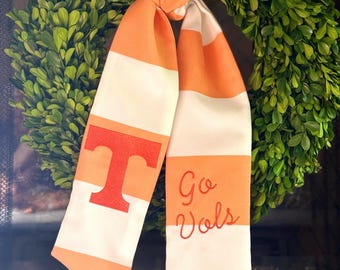Op de afbeelding: Een sjaal met witte en oranje strepen, met een oranje "T" en de geborduurde woorden "Go Vols". De sjaal is gedrapeerd over een groene krans.