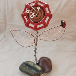 Könnte beinhalten: Eine rote Metallblume mit einem Holzknopf in der Mitte, umgeben von Drahtblättern und einem Marienkäfer. Die Blume steht auf zwei Steinen.