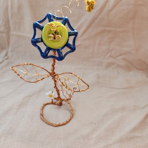 Könnte beinhalten: Eine Draht-Skulptur einer Blume mit einem blauen und grünen Knopf als Mitte. Die Blume hat einen Stiel und Blätter aus Kupferdraht. Die Skulptur steht auf einem weißen Hintergrund.