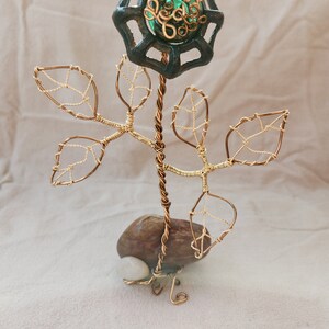 Könnte beinhalten: Eine Drahtskulptur einer Blume mit einem grünen Glaskern und goldenen Drahtblättern. Die Blume ist an einem braunen Stein mit einem weißen Stein an der Basis befestigt.