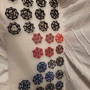 Pode incluir: Uma coleção de manípulos de válvulas de água vintage em várias cores, incluindo preto, vermelho e azul. Os manípulos estão dispostos numa superfície branca, mostrando os seus desenhos em forma de estrela. Ideal para decoração industrial.