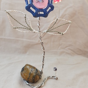Könnte beinhalten: Eine Drahtskulptur einer Blume mit einem blauen und rosafarbenen Knopf in der Mitte, einem rosafarbenen Schmetterling und grünen Drahtblättern. Die Blume steht auf einem braunen und schwarzen Stein.