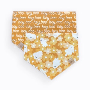 May include: Two triangular pet bandanas in mustard yellow. The top bandana features a repeating white script pattern reading hey, boo. The bottom bandana displays a white floral and ghost pattern with a small tan fabric tag on the corner.