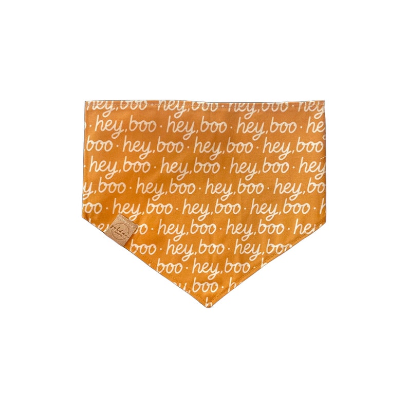 May include: A triangular pet bandana in a vibrant orange color featuring a repeating white script pattern that reads hey, boo. The fabric accessory includes a small, square-shaped tan leather-like brand tag attached to the lower left corner.