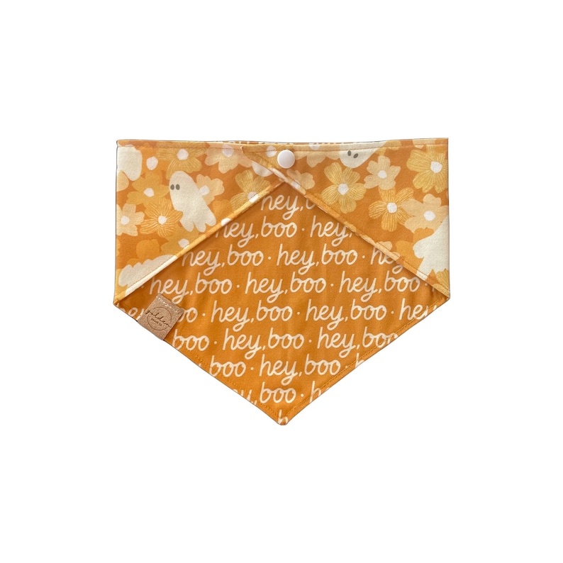 May include: A triangular pet bandana in mustard yellow featuring a repeating white text pattern that reads hey, boo. The fabric includes a floral and ghost print in cream and white tones, finished with a single white snap button closure.