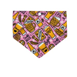 May include: Pink dog bandana with a repeating pattern of beer-related graphics in yellow and white. The design includes beer mugs, bottles, and cans, with text such as "ALE", "PORTER", and "Craft Beer". A white snap closure is at the top.