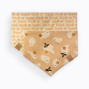 Puede incluir: Un pañuelo para mascotas con temática de Halloween con un fondo beige. El pañuelo presenta un patrón repetido de fantasmas con sombreros de bruja y sosteniendo calabazas. La capa superior tiene las palabras "trick or treat" en naranja.