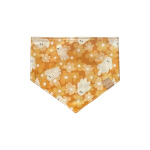 May include: A triangular pet bandana featuring a mustard yellow background with a repeating pattern of white flowers and small, friendly white ghosts with simple grey eyes. A small tan fabric tag is sewn onto the lower right corner edge.