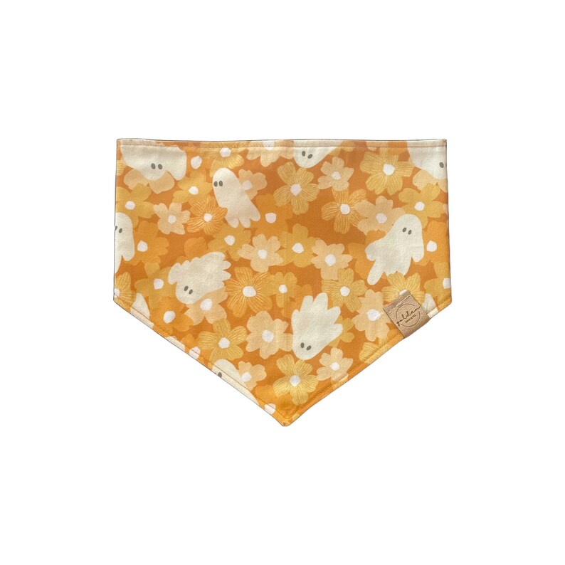 May include: A triangular pet bandana featuring a mustard yellow background with a repeating pattern of white flowers and small, friendly white ghosts with simple grey eyes. A small tan fabric tag is sewn onto the lower right corner edge.