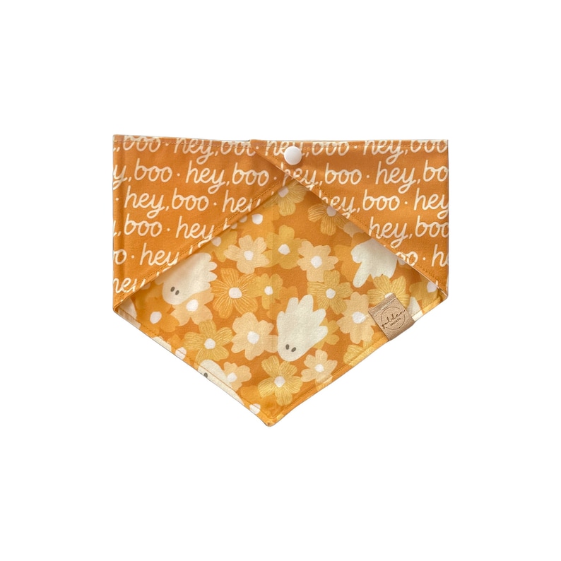 May include: A triangular pet bandana featuring a mustard yellow fabric with a repeating white text pattern reading hey boo. The reverse side displays a floral print in shades of orange and yellow with small white ghost figures and a small tan label.