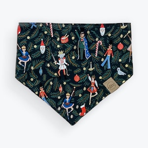 May include: A dark green dog bandana with a repeating pattern of Christmas-themed illustrations. The design includes nutcrackers, ballerinas, Santa faces, and ornaments. A small, round tag is attached to the bandana.