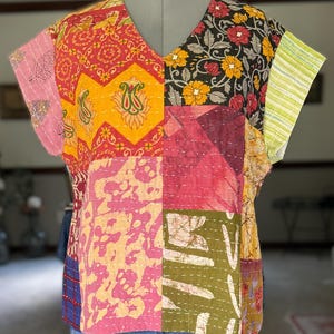 May include: A short-sleeved V-neck top constructed from various patchwork fabric panels featuring diverse patterns in shades of pink, yellow, orange, black, green, and blue, accented with visible white running stitch embroidery throughout the garment.