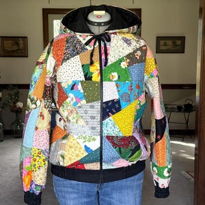 May include: A patchwork jacket with a black hood and zipper. The jacket is made of various fabric patterns in a variety of colors, including red, yellow, blue, and green. The jacket has a black drawstring and black cuffs.