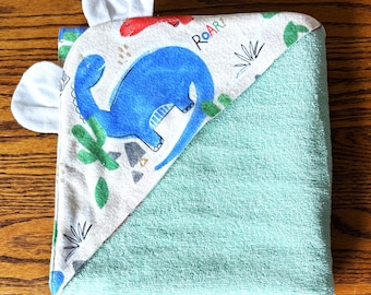 May include: A hooded baby towel with a dinosaur theme. The towel features a blue dinosaur illustration, the word "ROAR!", and white felt ears. The towel is light green and white with a dinosaur print.