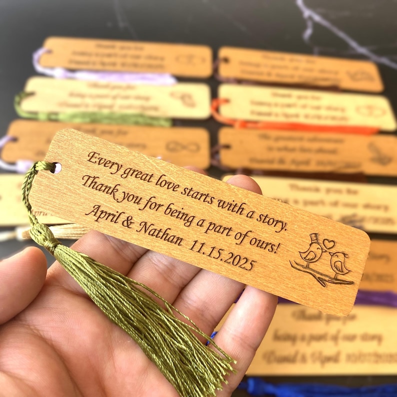 May include: Wooden bookmark with a green tassel, featuring the text "Every great love starts with a story. Thank you for being a part of ours! April & Nathan 11.15.2025" and a design of two birds with a heart.