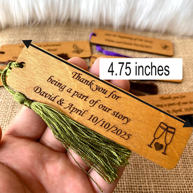 May include: Wooden bookmark with a green tassel, measuring 4.75 inches long. The bookmark is engraved with the text "Thank you for being a part of our story David & April 10/10/2025" and a design of two champagne glasses with a heart.