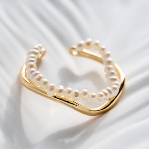 Puede incluir: Un brazalete de oreja dorado con un diseño de doble banda. La banda superior está adornada con pequeñas perlas redondas. El brazalete tiene un acabado liso y pulido y está sobre un fondo blanco.