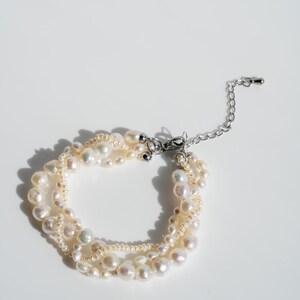 May include: A multi-strand pearl bracelet with a silver clasp and chain. The bracelet features various sizes of off-white pearls, creating a layered effect. The pearls have a natural luster and are arranged in a circular design.