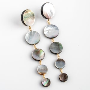 May include: Dangling earrings featuring iridescent, circular mother-of-pearl pendants. The earrings have a gold-colored chain connecting the pendants. The top pendant is a dark color, and the other pendants are a lighter color.